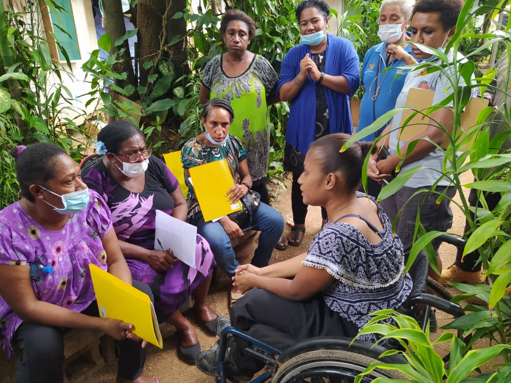 A woman sitting outside in a wheelchair talks with several female health personnel sitting and standing in front of her